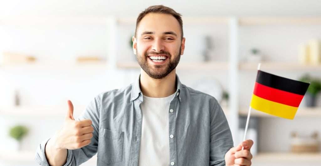 Happy Caucasian man showing thumb up and flag of Germany, posing and smiling at camera indoors